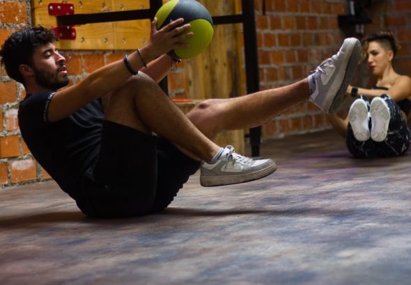 A man and woman perform core exercises with a medicine ball in a brick wall gym.